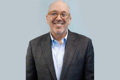 Headshot of Gregory D. Hess -- A white man stands in a grey suit with orange glasses against a light grey wall.