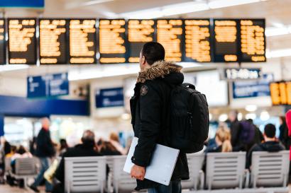 Man standing with backpack and laptop in hand looking at departure board at the airport.