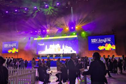 Large room with a presentation screen in the center, crowd dispersed throughout as festive lights flash.