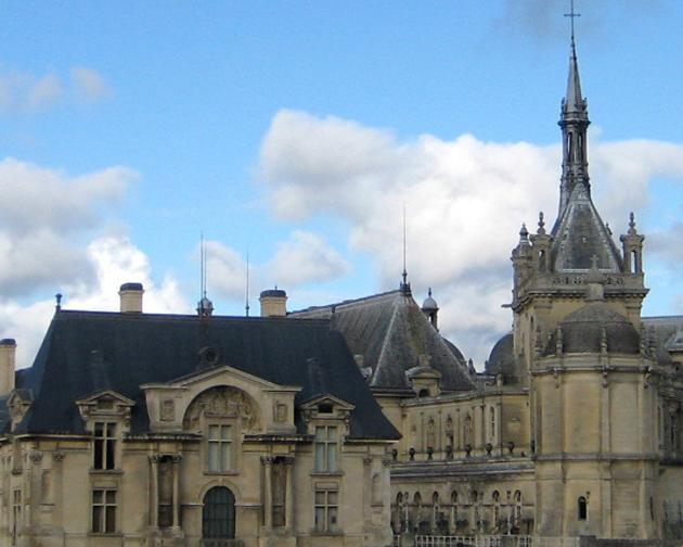 A European-style building with a cream exterior, blue roof, and multiple spires sits on a manicured lawn on a sunny day.