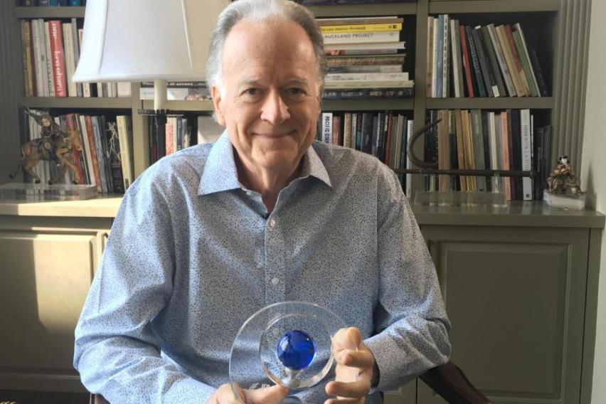 An older gentleman sitting holding a glass award. In the background is a lamp and bookshelf.