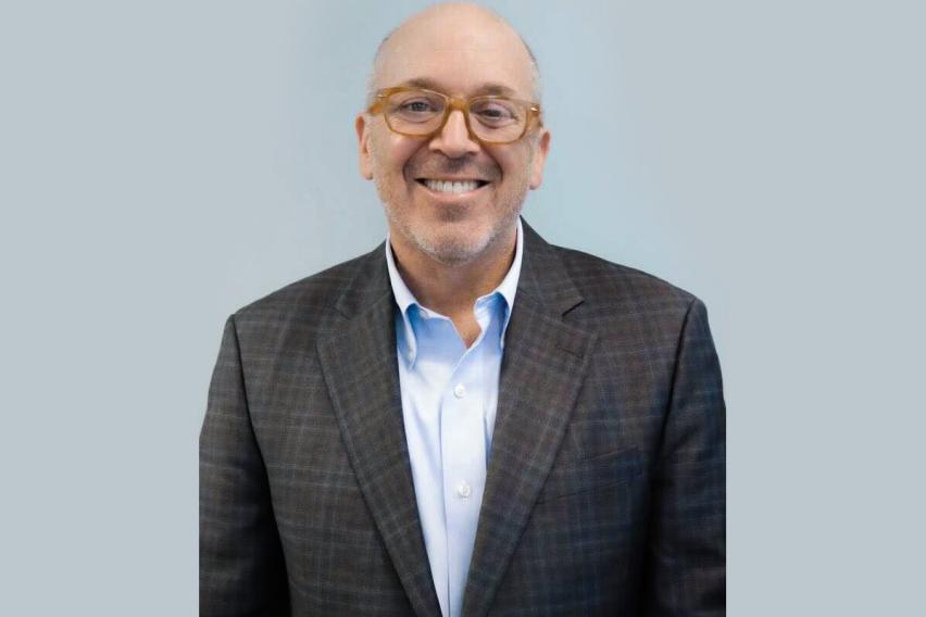 Headshot of Gregory D. Hess -- A white man stands in a grey suit with orange glasses against a light grey wall.