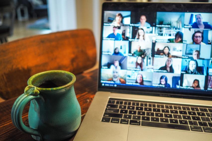 Coffee cup on a table next to a laptop with the gallery view of Zoom displaying dozens of participants.