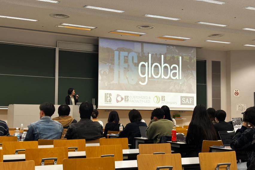 Natsuko Kambayashi presenting to a classroom at Hosei University, the screen featuring IES Global and its brands ie. IES Abroad, IES Customized, IES Internships, SAF. 