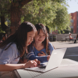 Two students sit outside at a table on their college campus working from a laptop.