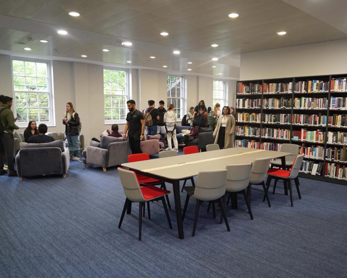 Library room with a conference table in front of bookshelves and various students in the sitting area.