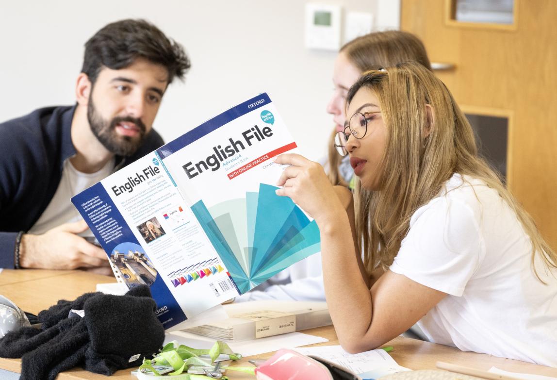 Three students in English class, one holding book that two are reading. 