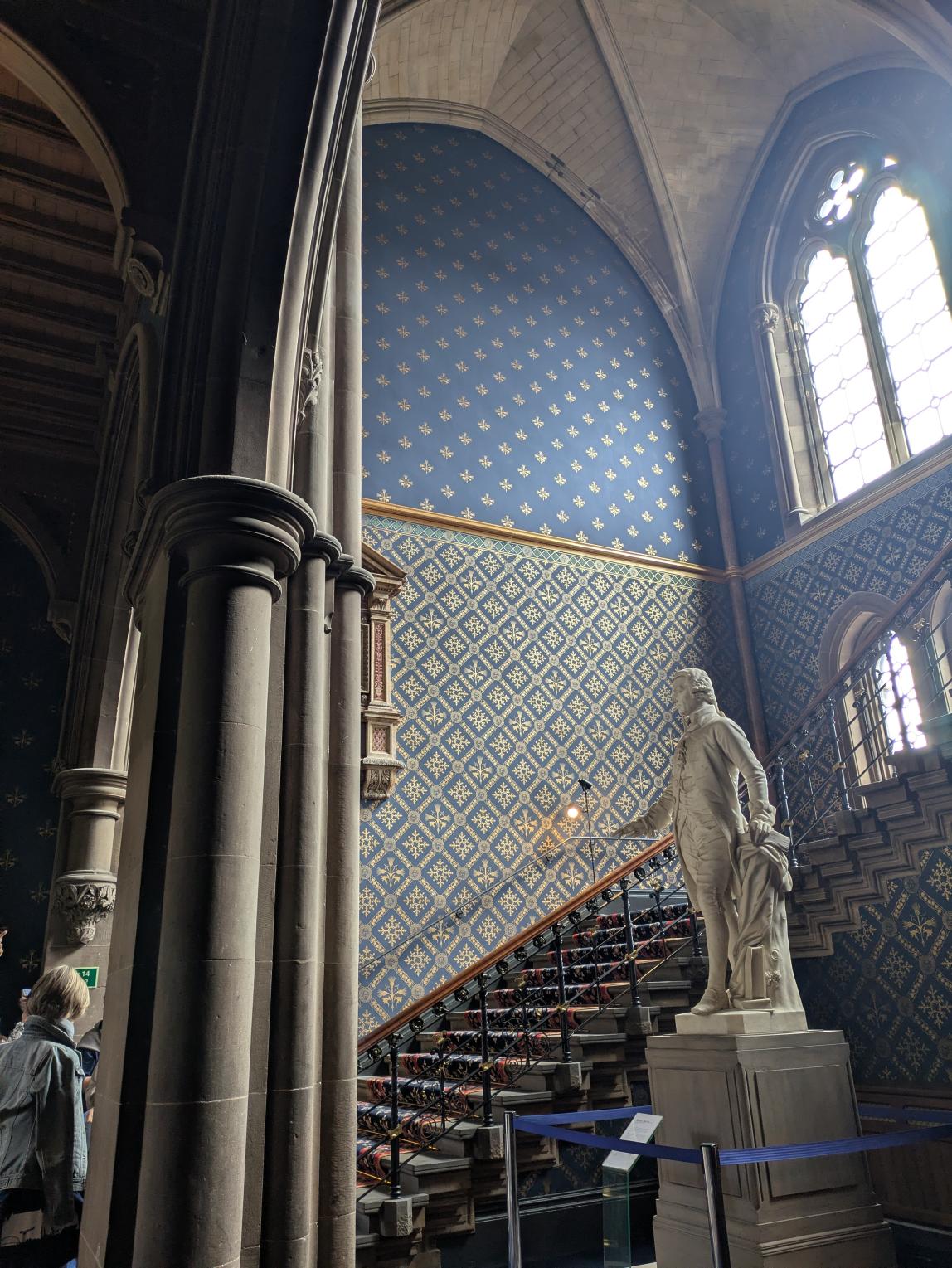 Regal chamber with a statue, high ceilings, ornate wallpaper and sunlight coming through a cathedral-like window.