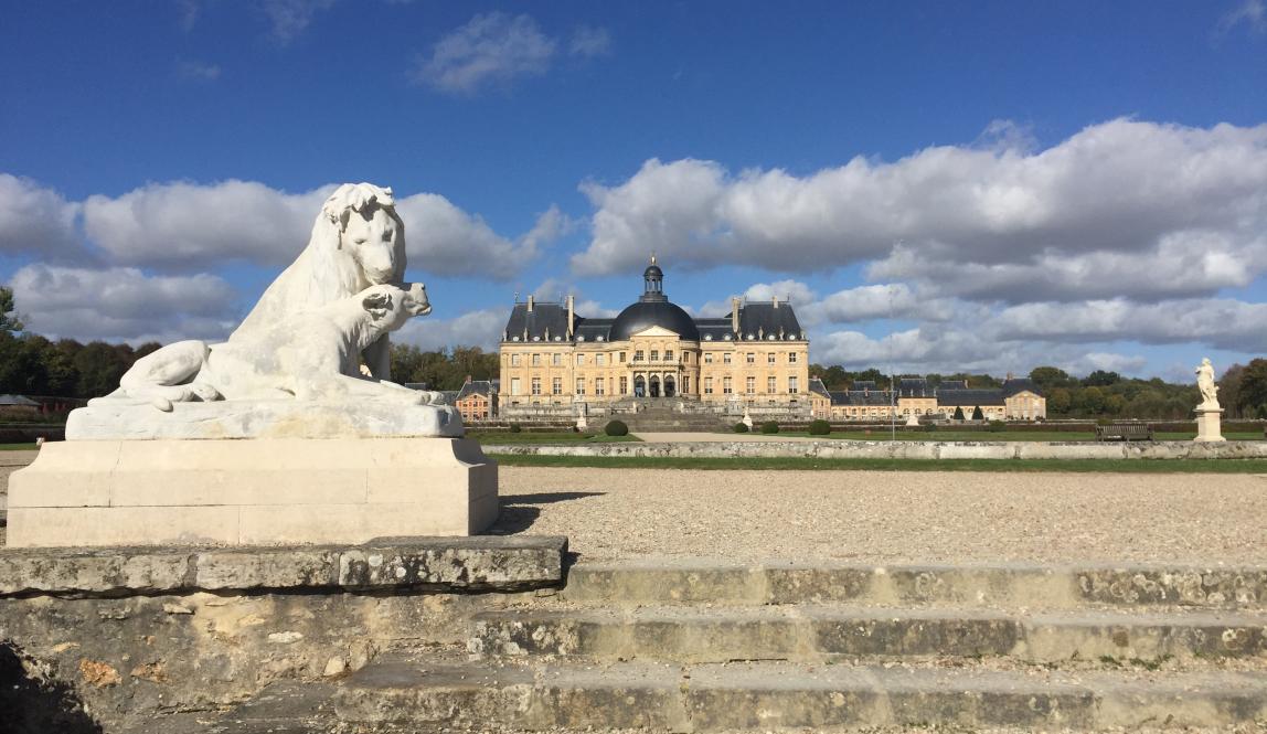 A statue of two lions sitting together in the foreground of the palace of Fontainebleau. A building with a cream exterior and dark roof, along with other statues, sit in the background.