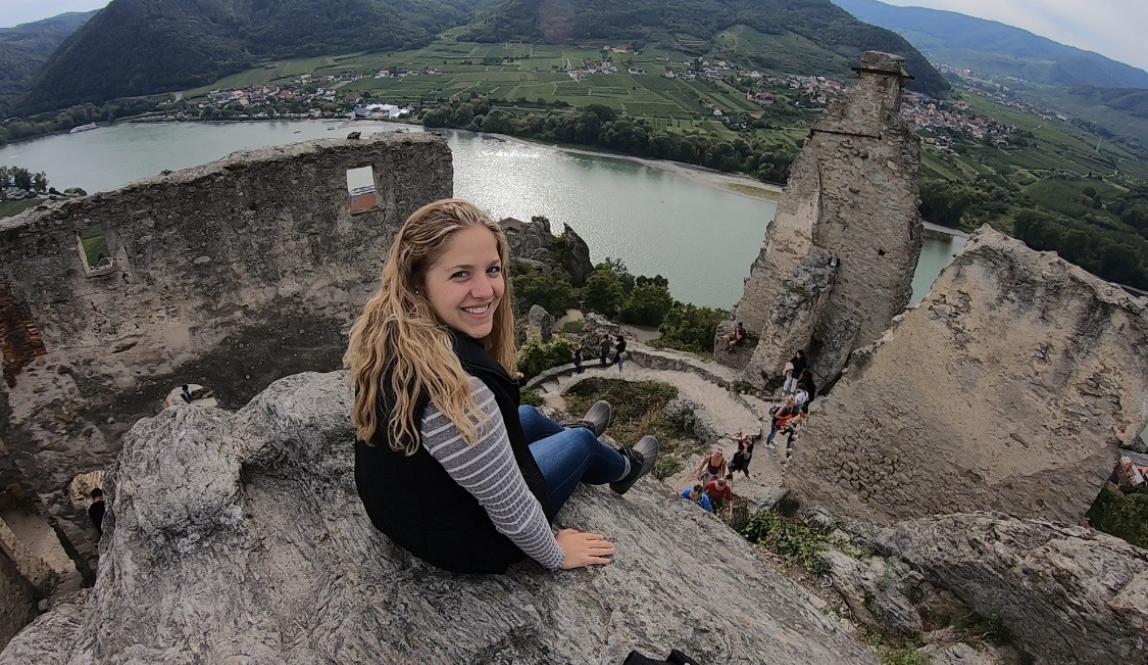 A student sitting atop Castle Ruins in Wachau. There is a river a few feet below, and hills and houses in the background landscape.