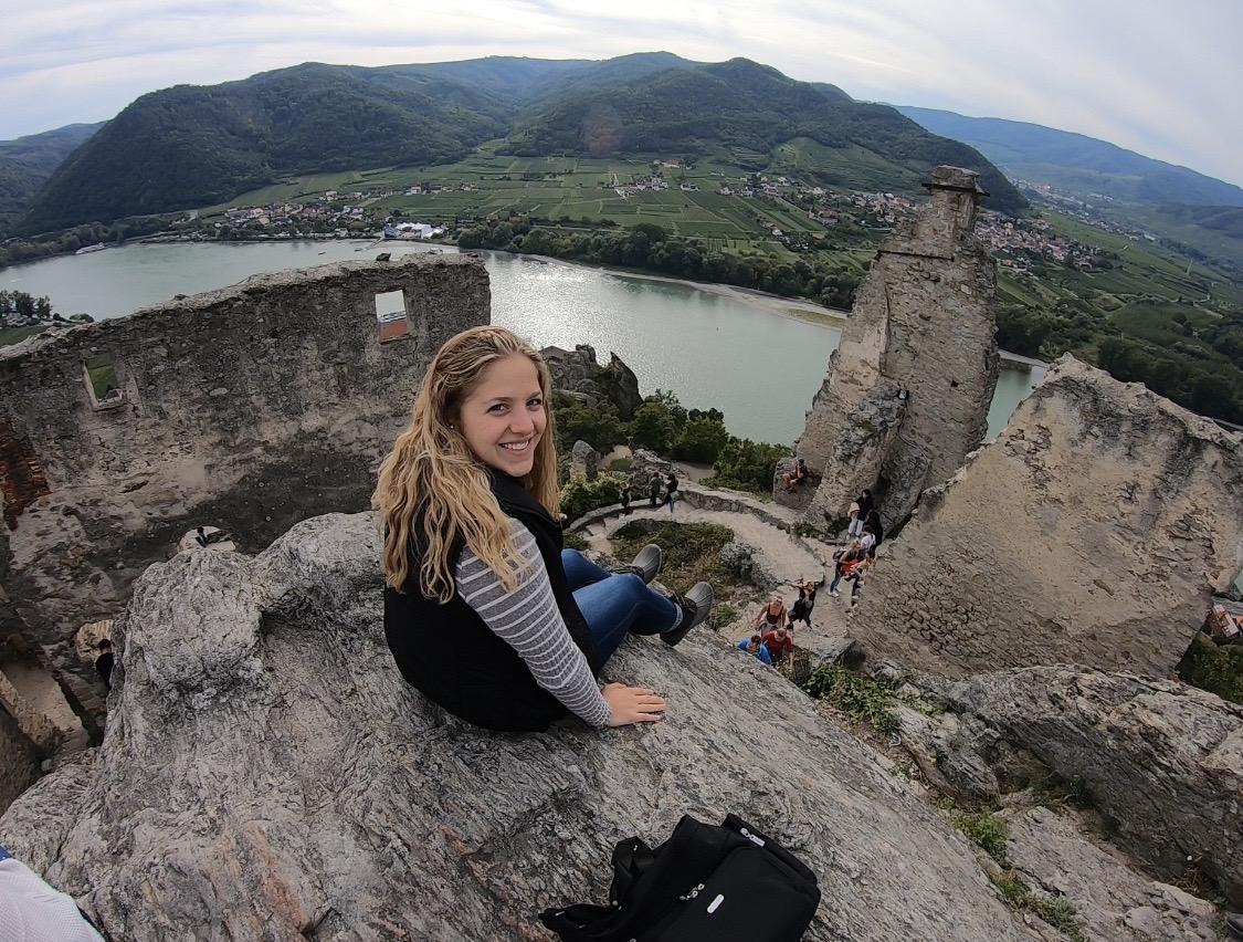 A student sitting atop Castle Ruins in Wachau. There is a river a few feet below, and hills and houses in the background landscape.