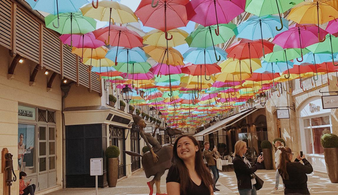 A student stands underneath The Umbrella Sky Project, an art display featuring hundreds of brightly colored umbrellas above the streets in Paris.