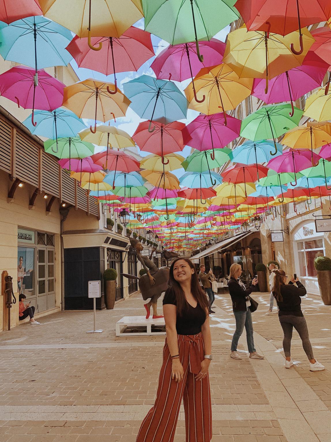 A student stands underneath The Umbrella Sky Project, an art display featuring hundreds of brightly colored umbrellas above the streets in Paris.