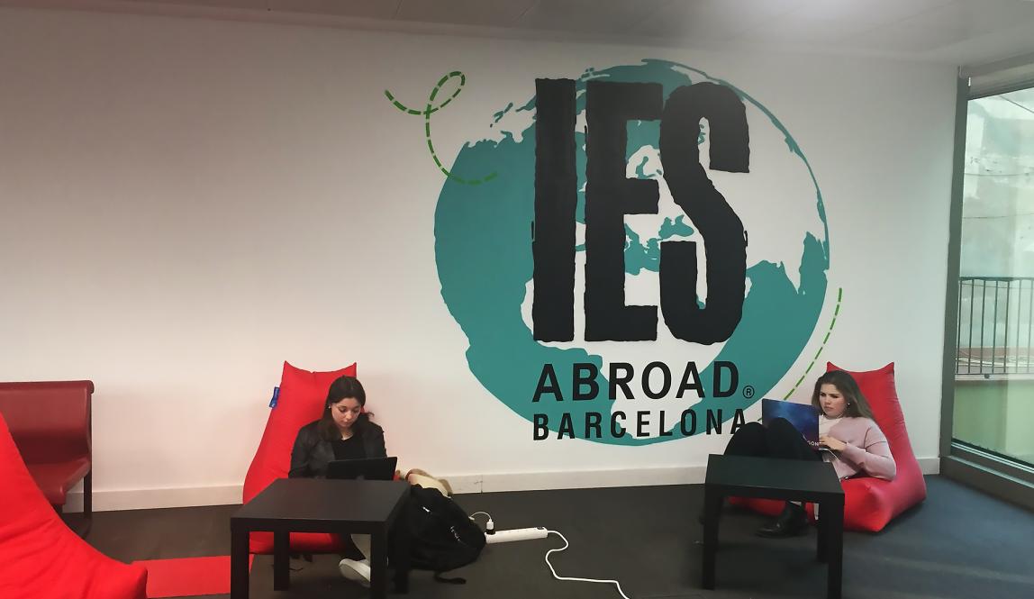 Two students sit in red lounge chairs in the Barcelona Center. Between them in a globe painted on the wall.