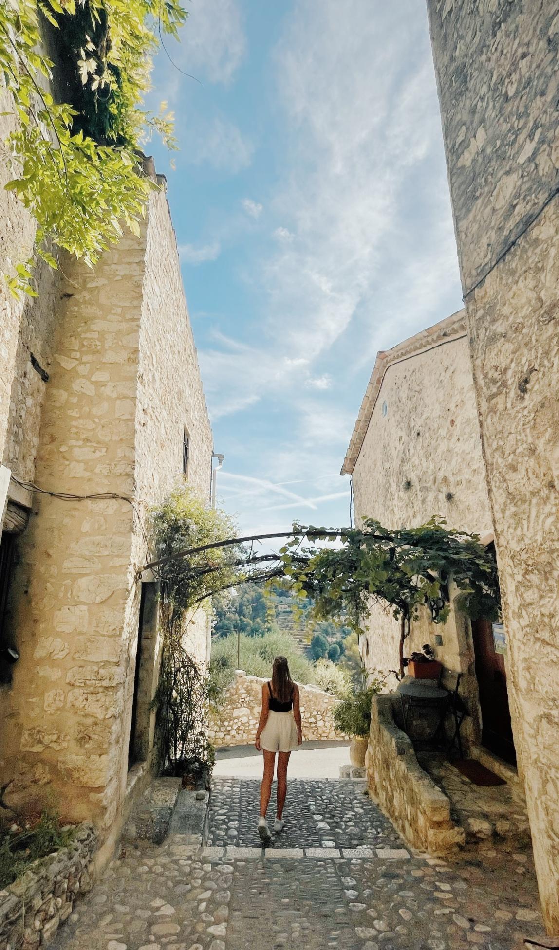 A students stands between two buildings in Nice, France. In front of her are trees.