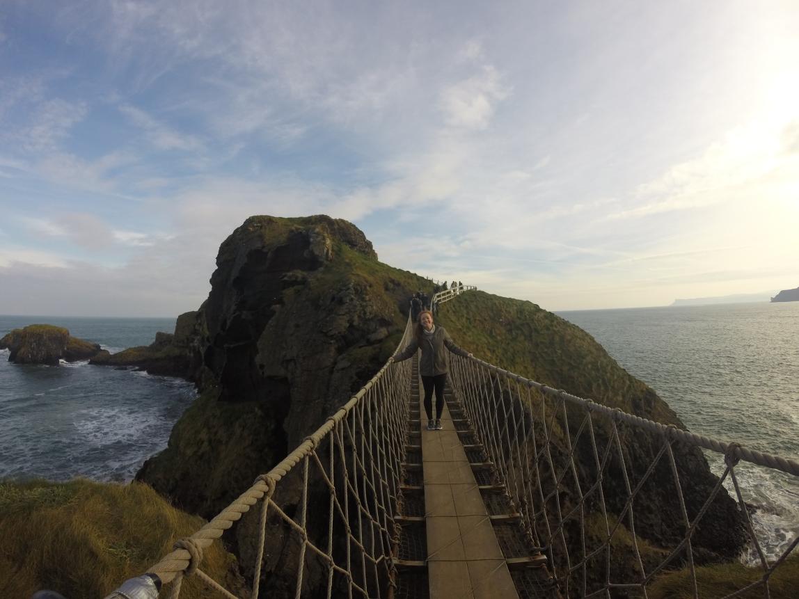 A student walks across a narrow swinging bridge over water.