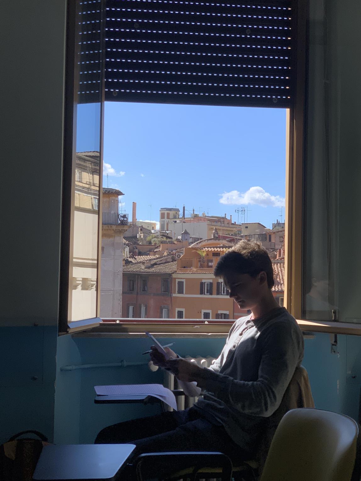 A student at the Rome Center works on their assignment with the window open. Outside the window are terracotta-colored buildings.