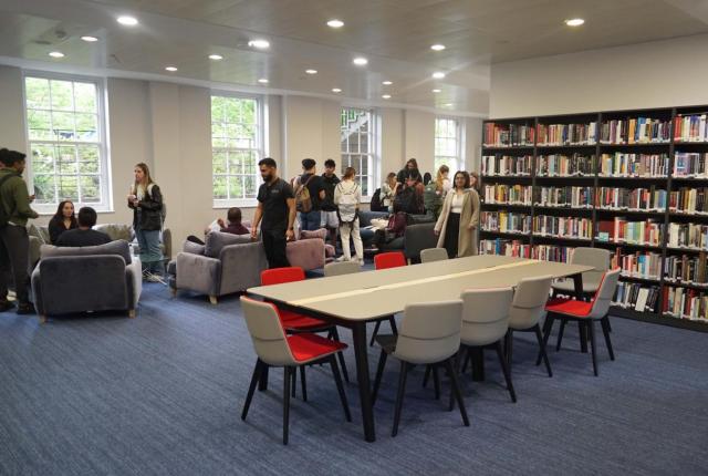 Library room with a conference table in front of bookshelves and various students in the sitting area.