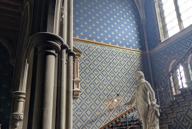 Regal chamber with a statue, high ceilings, ornate wallpaper and sunlight coming through a cathedral-like window.