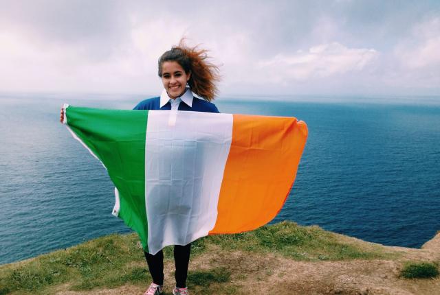 a student holding an Irish flag at the Cliffs of Moher