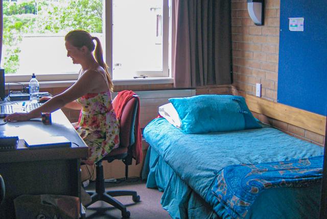 A student works at their desk in a small bedroom with a large window, a bed, and comfortable seating.