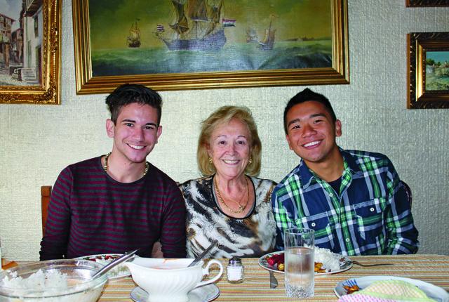 Students sit with their host mother at the dinner table, smiling at the camera.