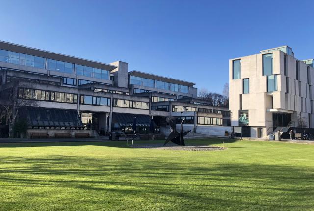 A sleek, modern, mostly glass building on Dublin Trinity College's manicured campus. There is a modern art statue made of black metal in front of the building.