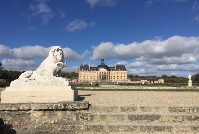 A statue of two lions sitting together in the foreground of the palace of Fontainebleau. A building with a cream exterior and dark roof, along with other statues, sit in the background.