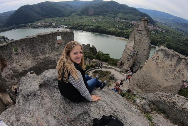 A student sitting atop Castle Ruins in Wachau. There is a river a few feet below, and hills and houses in the background landscape.