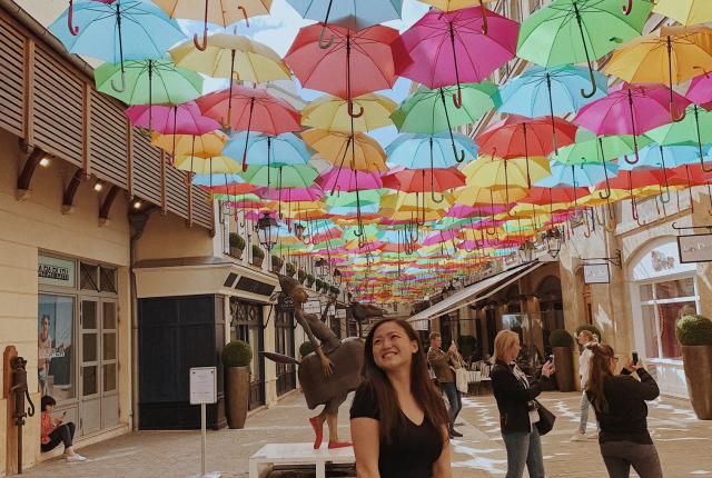 A student stands underneath The Umbrella Sky Project, an art display featuring hundreds of brightly colored umbrellas above the streets in Paris.