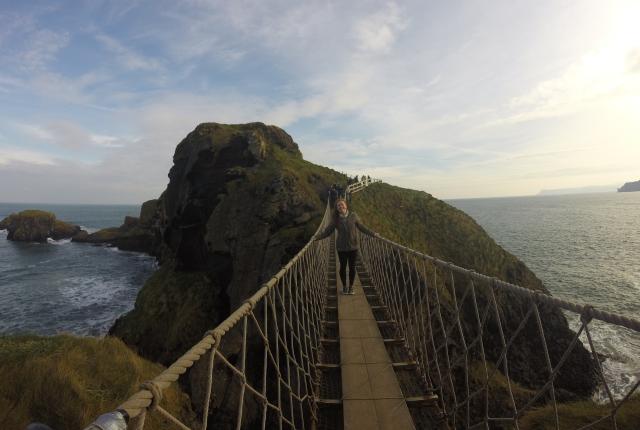 A student walks across a narrow swinging bridge over water.