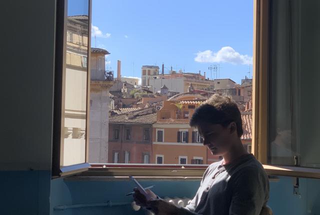 A student at the Rome Center works on their assignment with the window open. Outside the window are terracotta-colored buildings.