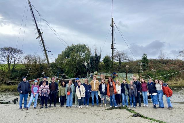 large group of students standing on a sand beach in front of a massive boat with grass, trees, and fog in the background