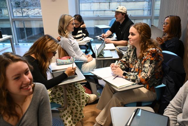 Several happy students are sitting in close proximity some have their desks turned around to face their classmates and a discussion is taking place