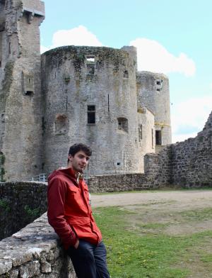 A student poses in front of ruins of an old, stone building on a sunny day.