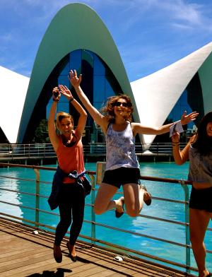 three students jumping on a boardwalk with clear blue water and a white building in the background