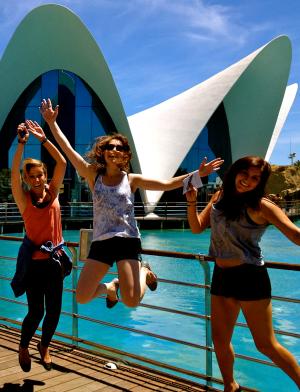 three students jumping on a boardwalk with clear blue water and a white building in the background