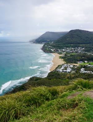 The coast in Royal National Park. Hills and greenery are further inland, leading to a clean, sandy beach and turquoise water.