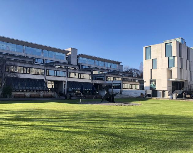 A sleek, modern, mostly glass building on Dublin Trinity College's manicured campus. There is a modern art statue made of black metal in front of the building.