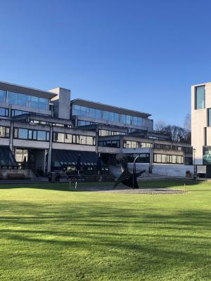 A sleek, modern, mostly glass building on Dublin Trinity College's manicured campus. There is a modern art statue made of black metal in front of the building.