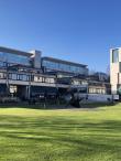 A sleek, modern, mostly glass building on Dublin Trinity College's manicured campus. There is a modern art statue made of black metal in front of the building.