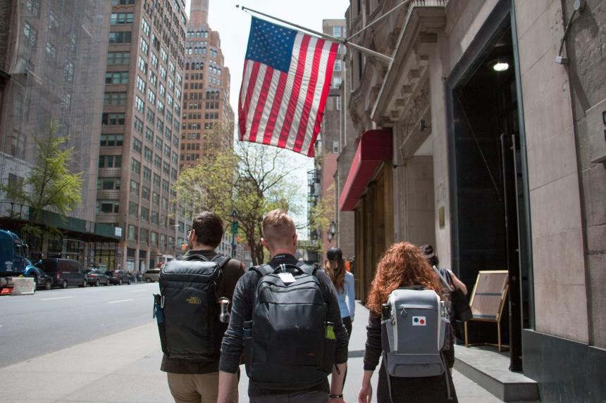 Three students wearing backpacks walk under an American flag in the U.S.