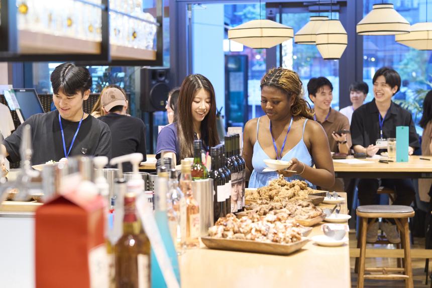 3 students grabbing food at counter, others chatting and laughing in the background.