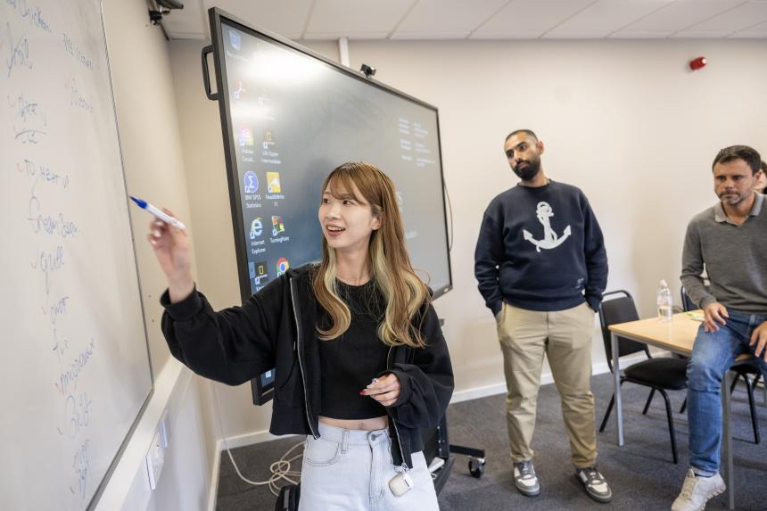 Student at a whiteboard during class, teacher and classmate behind her. 